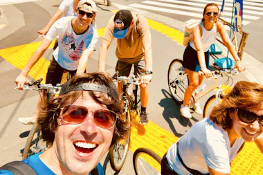 A group of five smiling people ride bicycles together on a sunny day, with one person taking a selfie in the front. They are on a city street near a crosswalk with bright yellow tactile paving.