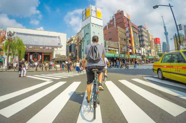 A person rides a bicycle across a busy crosswalk in an urban area, surrounded by pedestrians and buildings. A yellow taxi and Tokyo Skytree are visible in the background under a partly cloudy sky.