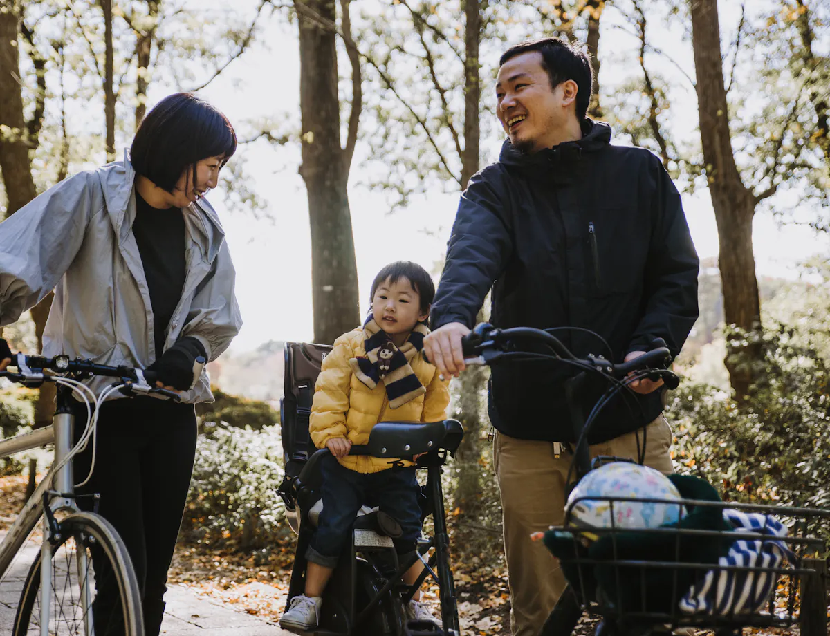 Cycling in Tokyo A woman and a man with bicycles smile at a young child sitting in a child bike seat. They are outdoors on a sunny day, surrounded by trees. The scene appears relaxed and cheerful.