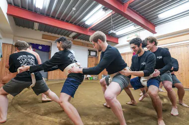 A group of people in casual athletic clothing participate in a sumo wrestling training exercise, lined up and holding each other's belts in a wooden dojo with a dirt floor.