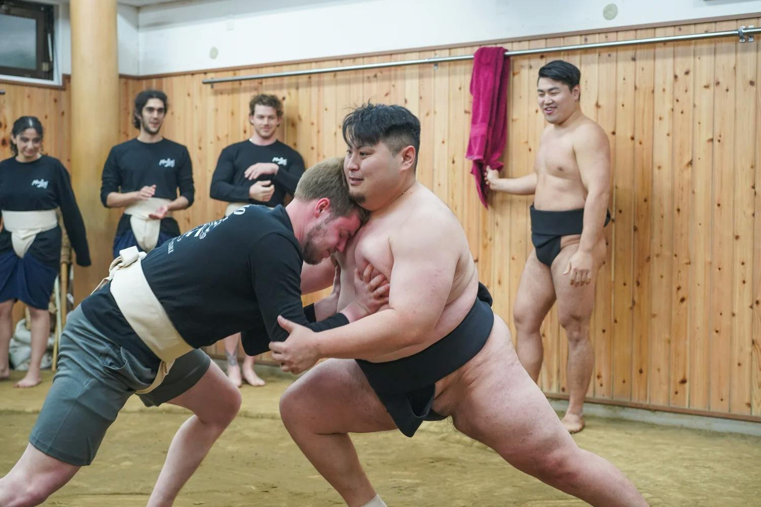 A sumo wrestler and a man in sportswear grapple in a wooden-paneled room, while others, including more sumo wrestlers and spectators, watch and smile in the background.