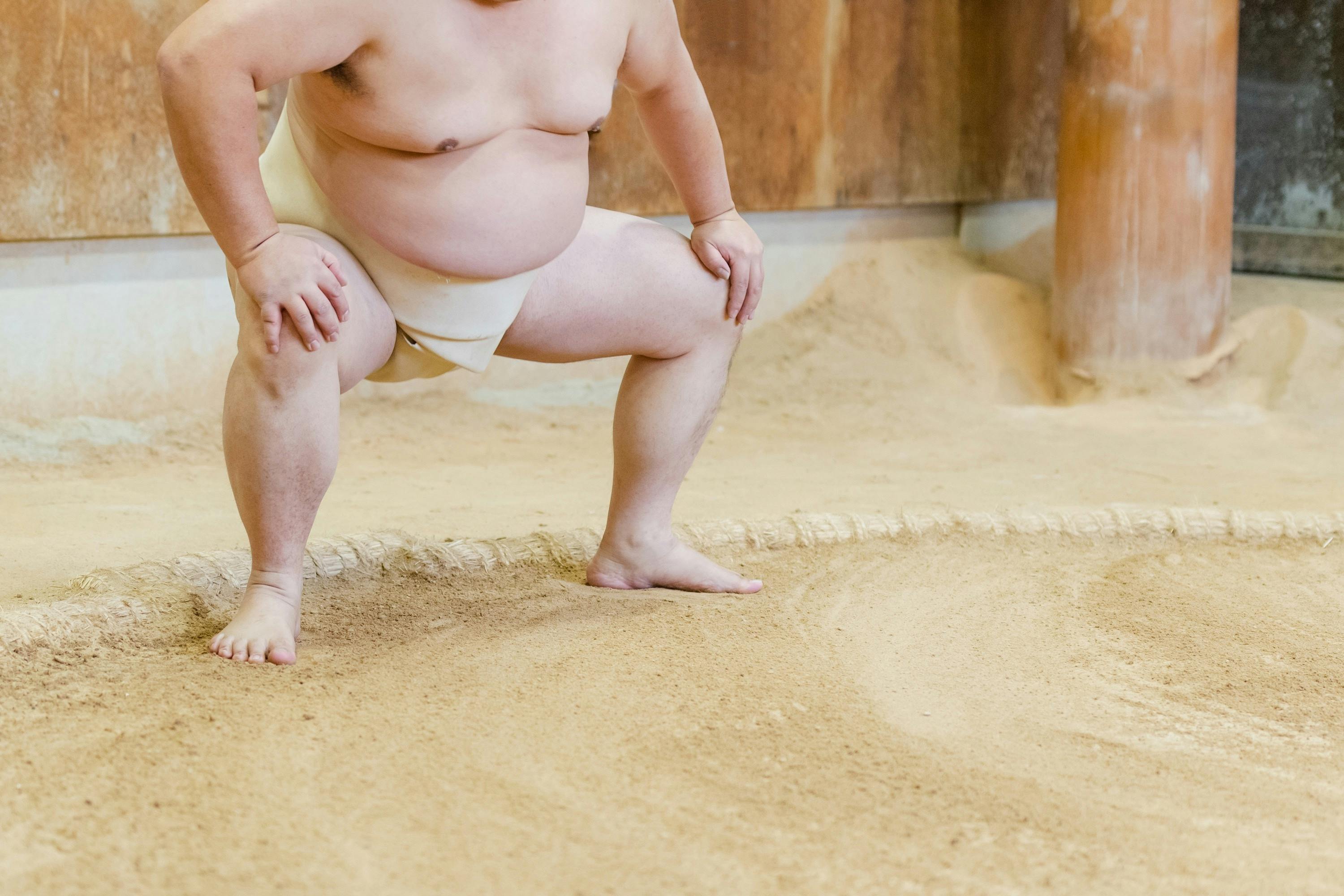 A sumo wrestler wearing a traditional mawashi squats in a sandy training ring, with hands on thighs, preparing for practice. The background shows wooden walls and a round sumo ring.