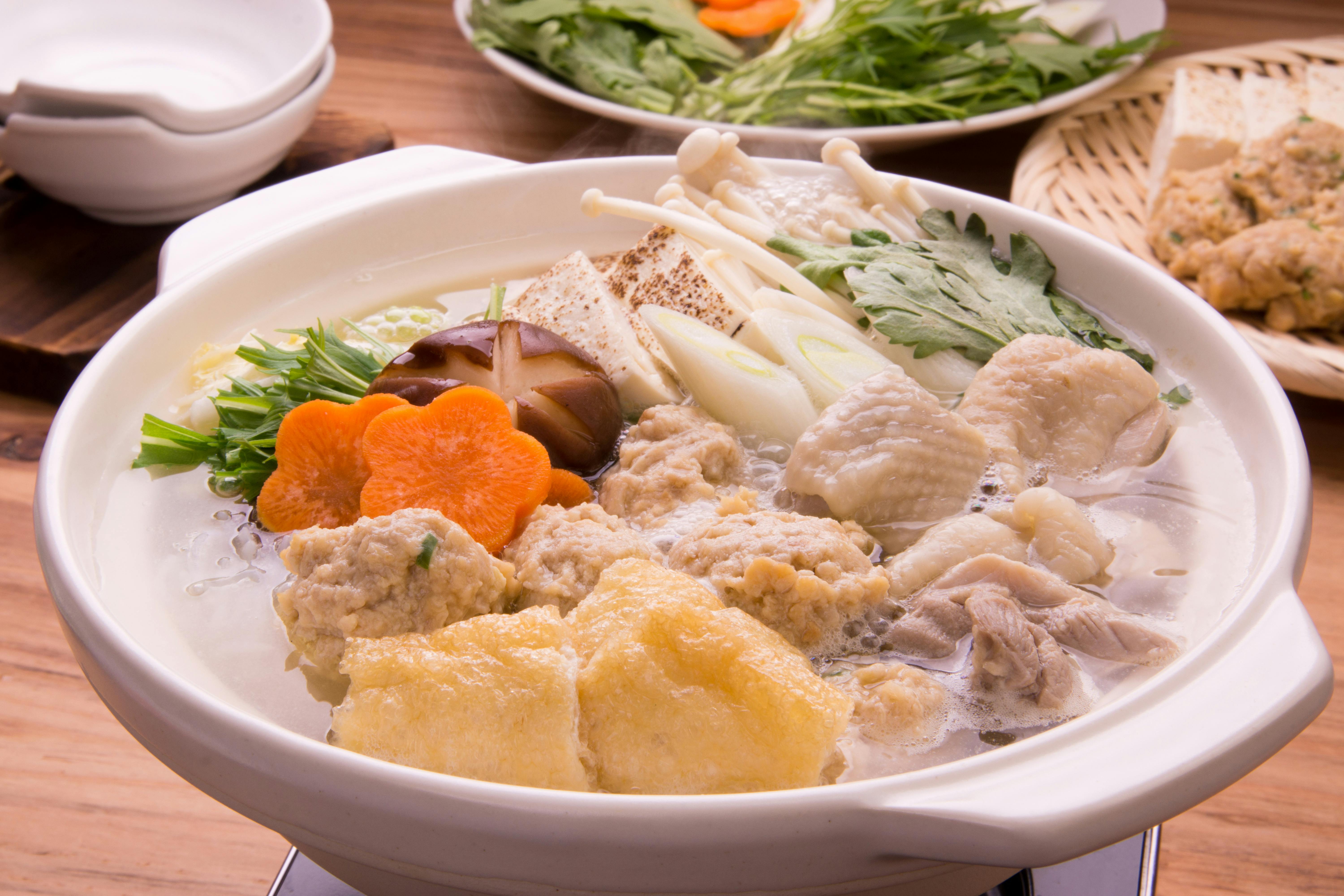 A hot pot filled with simmering ingredients including tofu, chicken, mushrooms, leafy greens, carrots, and fried tofu, with plates of raw vegetables and tableware in the background.