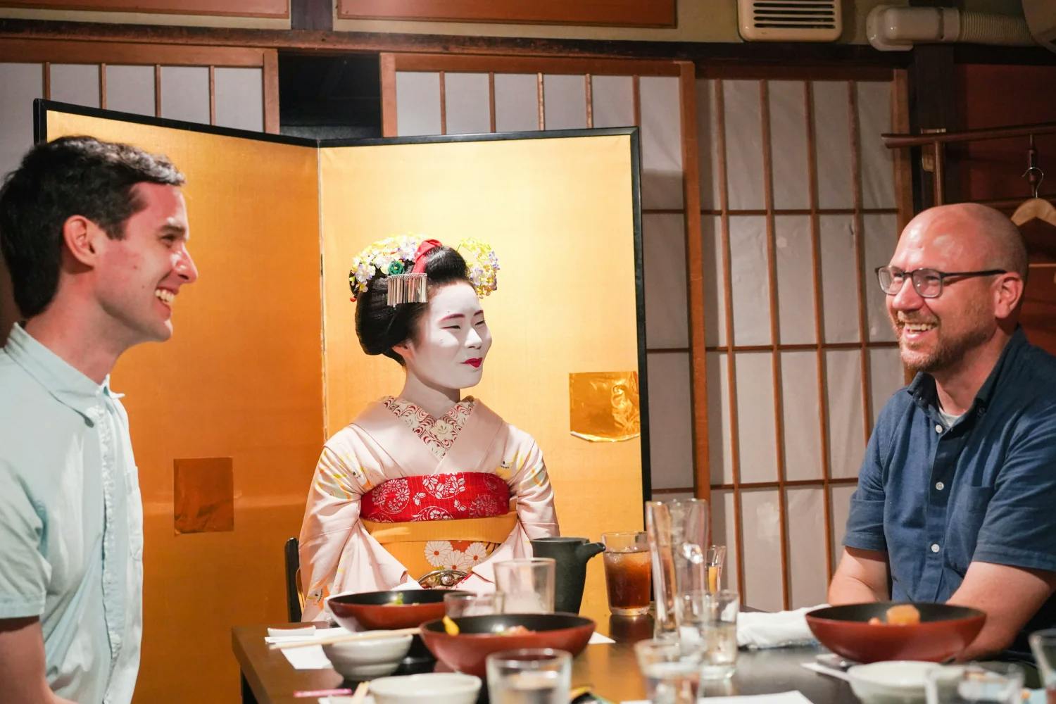 Two men smile and laugh while dining at a table, with a geisha in traditional attire seated between them. The setting appears to be a Japanese restaurant with wooden paneling and decorative screens.
