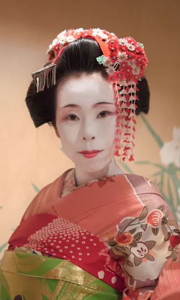 Maiko A woman dressed as a geisha wearing a colorful kimono with floral patterns, elaborate hair ornaments, and traditional white makeup, standing in front of a background with painted white flowers and green leaves.