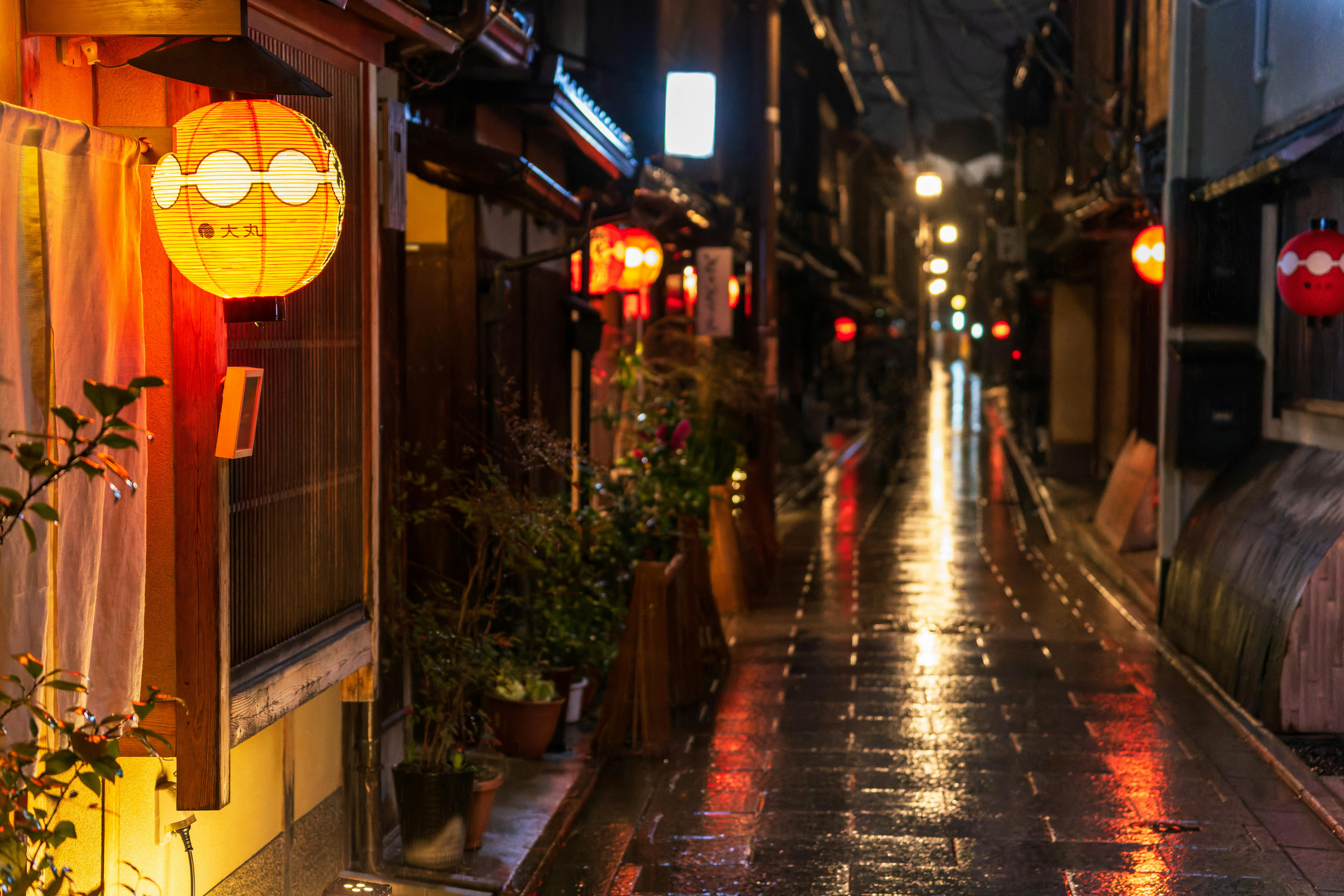 A narrow, rain-soaked alley in Japan glows with reflections and the warm light of red paper lanterns hanging outside traditional wooden buildings at night.