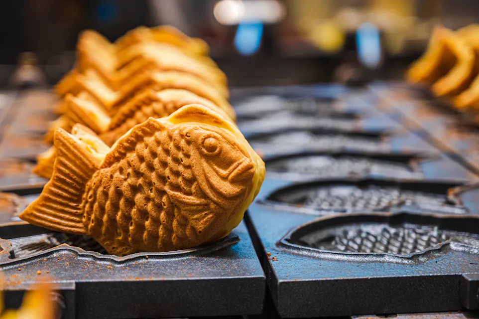 Taiyaki Close-up of rows of freshly baked fish-shaped pastries on a mold. The pastries are golden brown with textured scales and fins, arranged in a line, with a blurred background of more molds.