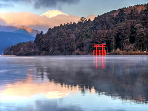 Lake Ashinoko A serene lake with misty waters reflecting surrounding trees and mountains. A bright red torii gate stands near the water's edge, and the snow-capped peak of Mount Fuji is visible in the background, partially obscured by clouds.