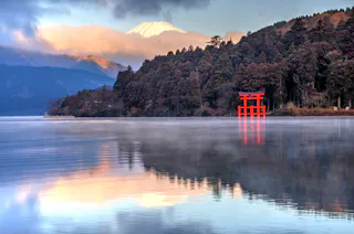 A serene lake with misty waters reflecting surrounding trees and mountains. A bright red torii gate stands near the water's edge, and the snow-capped peak of Mount Fuji is visible in the background, partially obscured by clouds.