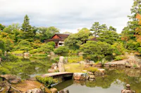 Traditional Japanese garden with a pond, stone pathway, lush greenery, rocks, and a wooden building surrounded by trees under a cloudy sky.