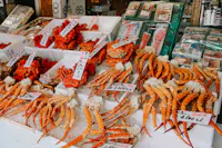 Market stall displaying fresh seafood, including red crab legs and other shellfish, with handwritten price tags in Japanese on white trays. Packaged seafood products are visible in the background.