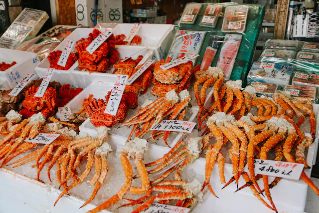 Market stall displaying fresh seafood, including red crab legs and other shellfish, with handwritten price tags in Japanese on white trays. Packaged seafood products are visible in the background.