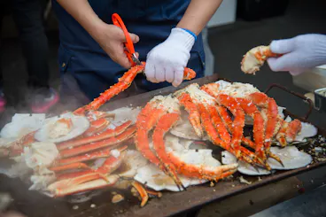 Two people wearing gloves prepare and cut large orange-red crab legs on a hot grill, surrounded by crab shells and steam rising from the seafood.