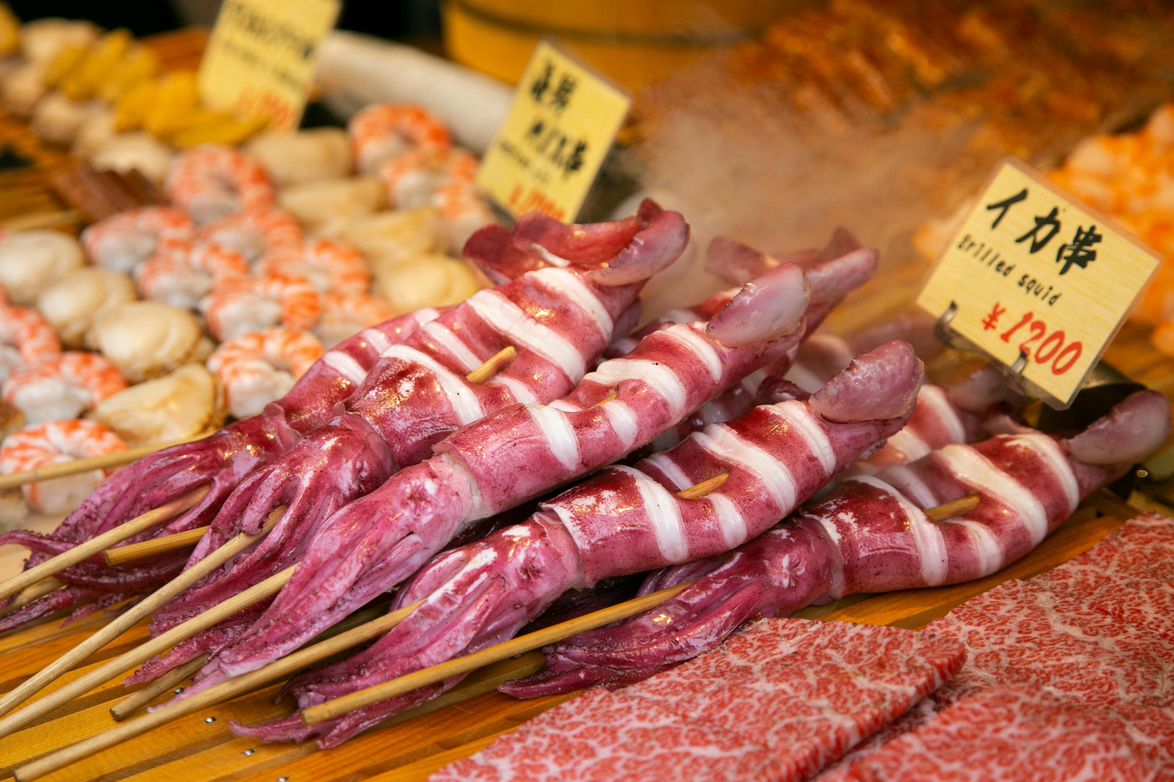 Skewered whole grilled squid are displayed on a wooden counter at a market, with a sign showing the price in yen. Other seafood items are visible in the background.