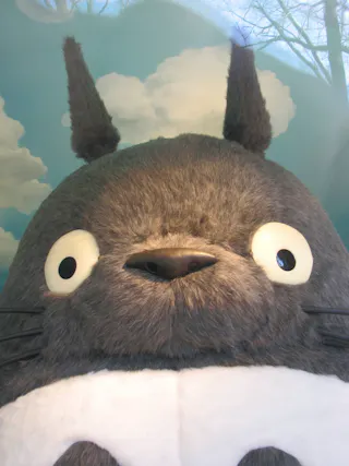 A close-up of a large, fluffy gray character with big, round eyes, pointy ears, and a white belly, set against a backdrop of blue sky and clouds.