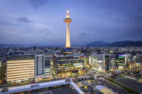 Kyoto Tower illuminated at dusk, standing above modern city buildings with mountains in the distance under a cloudy blue sky. Streets and buildings are lit up, creating a vibrant urban scene.
