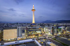 Kyoto Tower illuminated at dusk, standing above modern city buildings with mountains in the distance under a cloudy blue sky. Streets and buildings are lit up, creating a vibrant urban scene.