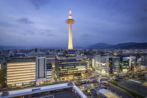 Kyoto Tower illuminated at dusk, standing above modern city buildings with mountains in the distance under a cloudy blue sky. Streets and buildings are lit up, creating a vibrant urban scene.