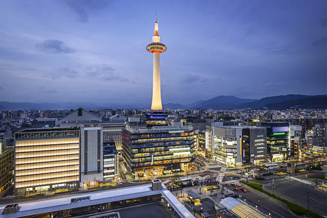 Kyoto Tower illuminated at dusk, standing above modern city buildings with mountains in the distance under a cloudy blue sky. Streets and buildings are lit up, creating a vibrant urban scene.