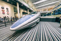 A futuristic bullet train on display inside a modern museum, with visitors observing and striped flooring beneath the train. Signs and another train are visible in the background.