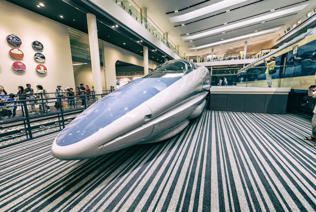 A futuristic bullet train on display inside a modern museum, with visitors observing and striped flooring beneath the train. Signs and another train are visible in the background.