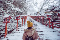 A person wearing a yellow hat and beige coat walks up a snow-covered stone staircase lined with red lanterns in a wintery Japanese shrine setting. Snow covers the trees and railings on both sides of the path.