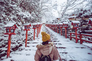A person wearing a yellow hat and beige coat walks up a snow-covered stone staircase lined with red lanterns in a wintery Japanese shrine setting. Snow covers the trees and railings on both sides of the path.