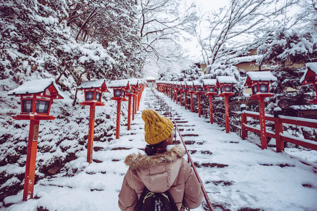 A person wearing a yellow hat and beige coat walks up a snow-covered stone staircase lined with red lanterns in a wintery Japanese shrine setting. Snow covers the trees and railings on both sides of the path.