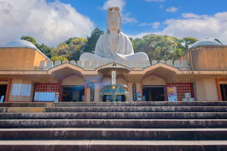 A large white statue of Buddha sits atop a temple structure, viewed from stone steps below, with incense burning in the foreground and green trees and blue sky in the background.