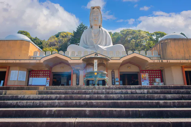 A large white statue of Buddha sits atop a temple structure, viewed from stone steps below, with incense burning in the foreground and green trees and blue sky in the background.