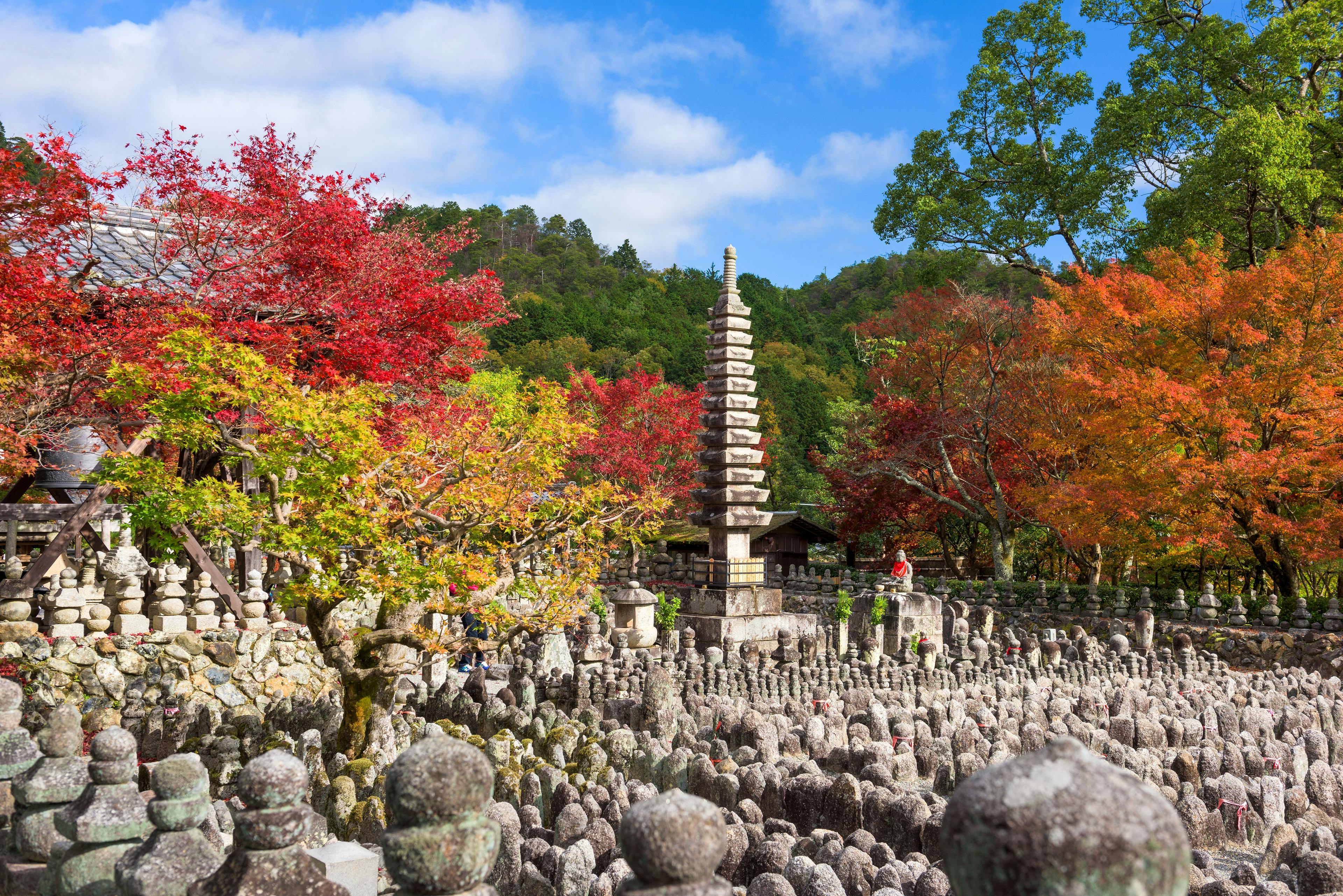 Stone pagoda and numerous stone statues surrounded by colorful autumn trees with red and orange leaves, set against a backdrop of green hills and a blue sky with scattered clouds.