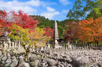 Stone pagoda and numerous stone statues surrounded by colorful autumn trees with red and orange leaves, set against a backdrop of green hills and a blue sky with scattered clouds.