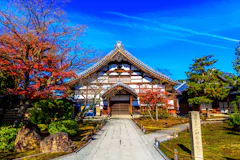 A traditional Japanese temple with a curved roof stands amid autumn trees with red and green foliage, under a clear blue sky with wispy clouds. A stone path leads to the entrance.