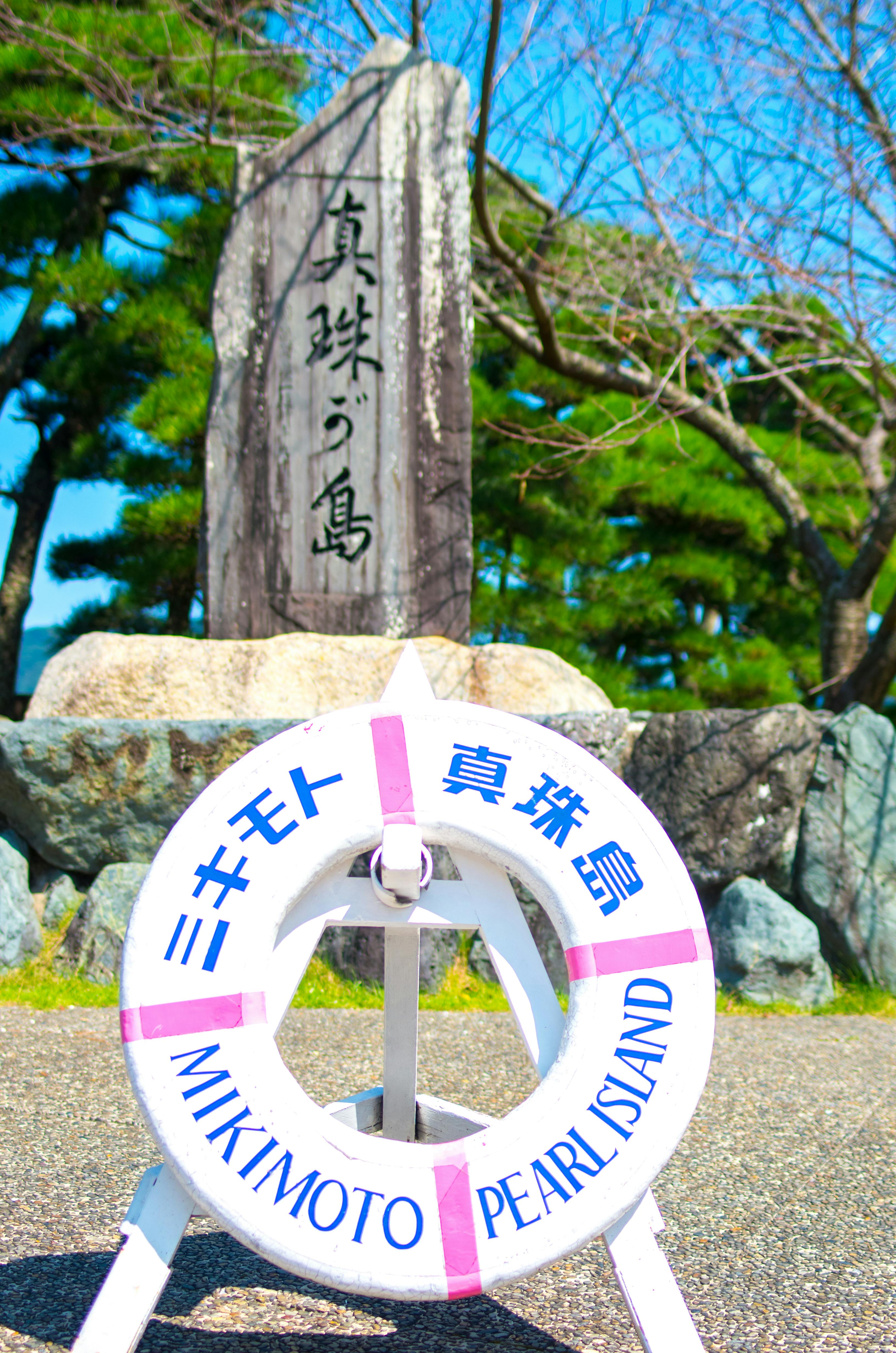 A white life preserver labeled "Mikimoto Pearl Island" in English and Japanese is displayed in front of a stone monument with Japanese inscriptions, surrounded by rocks, trees, and a clear blue sky in the background.