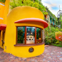 Ghibli Museum A large Totoro figure sits inside a curved yellow building with a big window, surrounded by lush greenery and colorful architecture at the Ghibli Museum in Japan.