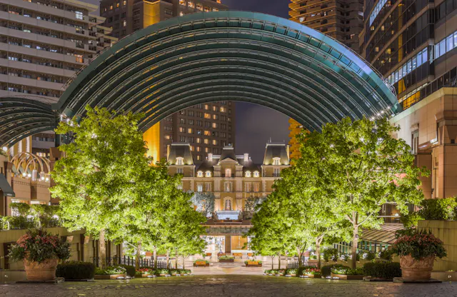 A brightly lit urban plaza at night, featuring green trees, a large arched glass canopy, and a classical-style building with a mansard roof, surrounded by tall modern buildings.