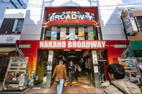 Entrance of Nakano Broadway shopping mall in Tokyo, Japan, with a red and white sign above and people entering and exiting. Vending machines and colorful advertisements line both sides of the entrance.