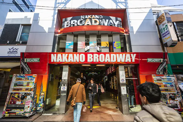 Entrance of Nakano Broadway shopping mall in Tokyo, Japan, with a red and white sign above and people entering and exiting. Vending machines and colorful advertisements line both sides of the entrance.