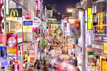 A bustling city street at night with vibrant signs and shopfronts, including McDonald's. Bright lights illuminate the crowded scene of people walking, creating a lively atmosphere. Tall buildings and colorful advertisements are visible in the background.