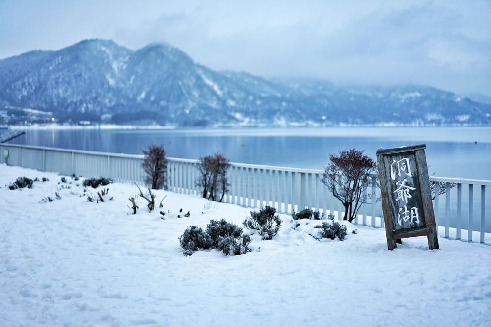 A lakeside scene in winter shows a snow-covered ground, sparse shrubs, a fence, and distant mountains. A standing sign with Japanese writing is displayed near the railing. The lake is calm, reflecting the overcast sky.
