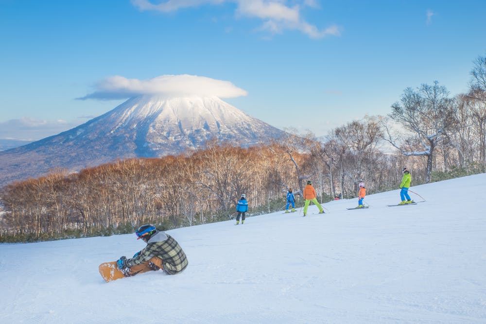 Several people ski and snowboard on a snowy slope with bare trees, while a snowboarder sits in the foreground. In the background, a snow-capped mountain with a cloud atop is visible under a blue sky.