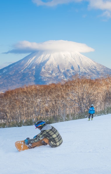 Niseko Ski Area Several people ski and snowboard on a snowy slope with bare trees, while a snowboarder sits in the foreground. In the background, a snow-capped mountain with a cloud atop is visible under a blue sky.
