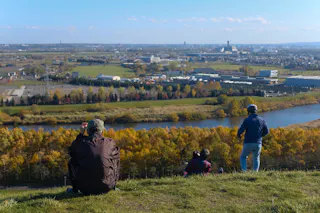 Three people sit and stand on a grassy hill overlooking a river, trees with autumn foliage, and a cityscape in the distance under a clear blue sky.
