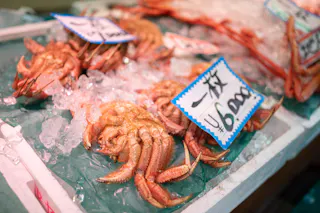 Cooked crabs displayed on ice in a seafood market, with handwritten price tags in Japanese placed above them. The crabs are arranged in rows on a green and white tray.