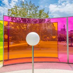 Kanazawa 21st Century Museum of Contemporary Art A white sphere stands in front of curved panels in yellow, orange, and pink, creating colorful reflections in an outdoor park with trees, grass, and modern buildings under a blue sky with clouds.
