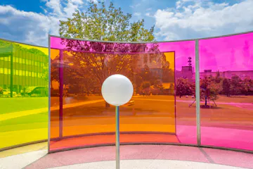 Kanazawa 21st Century Museum of Contemporary Art A white sphere stands in front of curved panels in yellow, orange, and pink, creating colorful reflections in an outdoor park with trees, grass, and modern buildings under a blue sky with clouds.