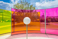 A white sphere stands in front of curved panels in yellow, orange, and pink, creating colorful reflections in an outdoor park with trees, grass, and modern buildings under a blue sky with clouds.