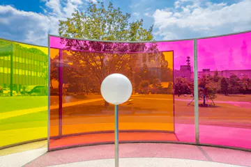 A white sphere stands in front of curved panels in yellow, orange, and pink, creating colorful reflections in an outdoor park with trees, grass, and modern buildings under a blue sky with clouds.