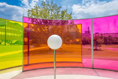 A white sphere stands in front of curved panels in yellow, orange, and pink, creating colorful reflections in an outdoor park with trees, grass, and modern buildings under a blue sky with clouds.