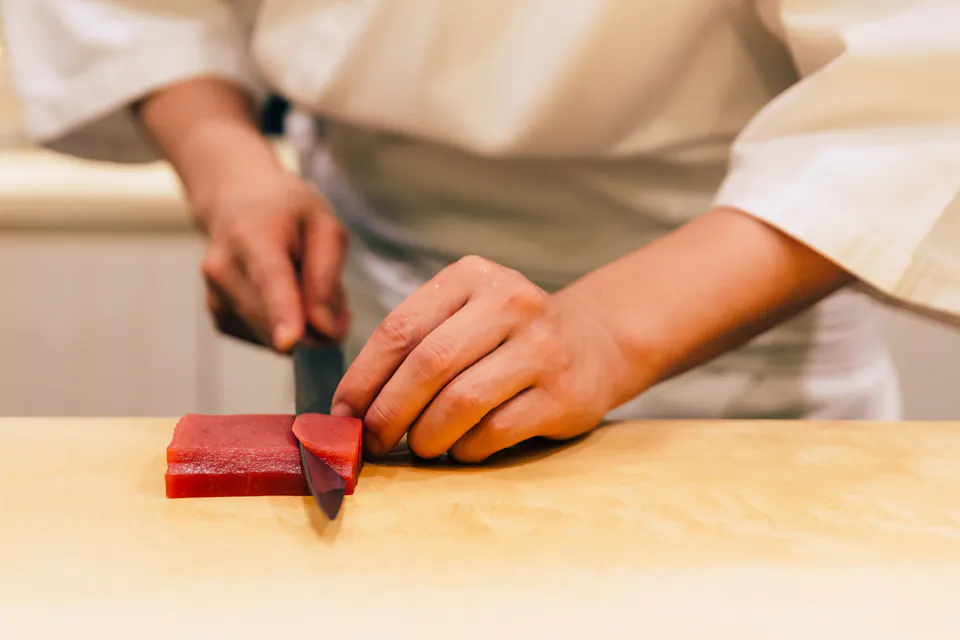 A chef in a white uniform carefully slices a piece of raw tuna on a light wooden cutting board. The chef's hands and knife are prominently shown, focusing on the precision of the slicing technique.